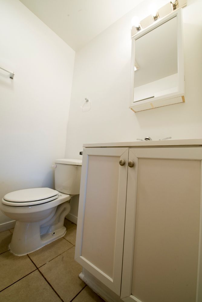 Small white bathroom with toilet, vanity, and mirror. Tan tiled floor.