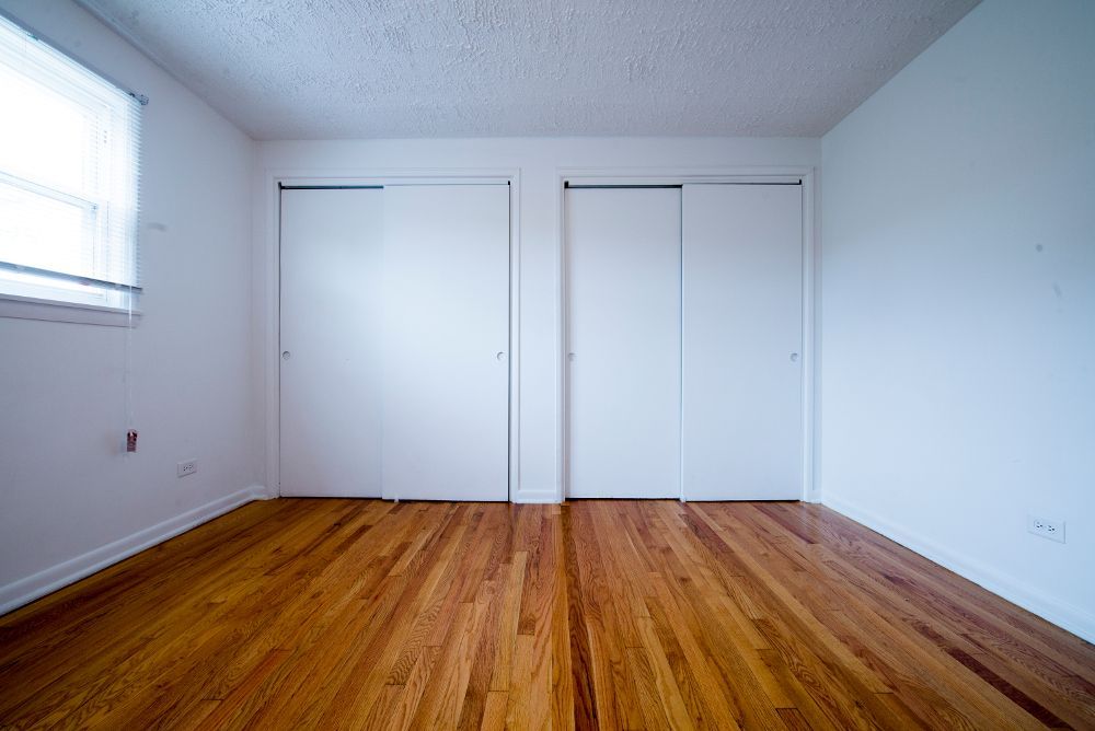 Empty bedroom with hardwood floor, two white closet doors, and a window on the left.