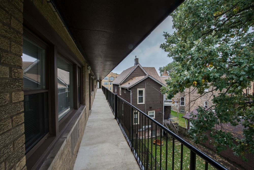Exterior view of a narrow balcony with a metal railing, overlooking a residential neighborhood with brick buildings and trees.