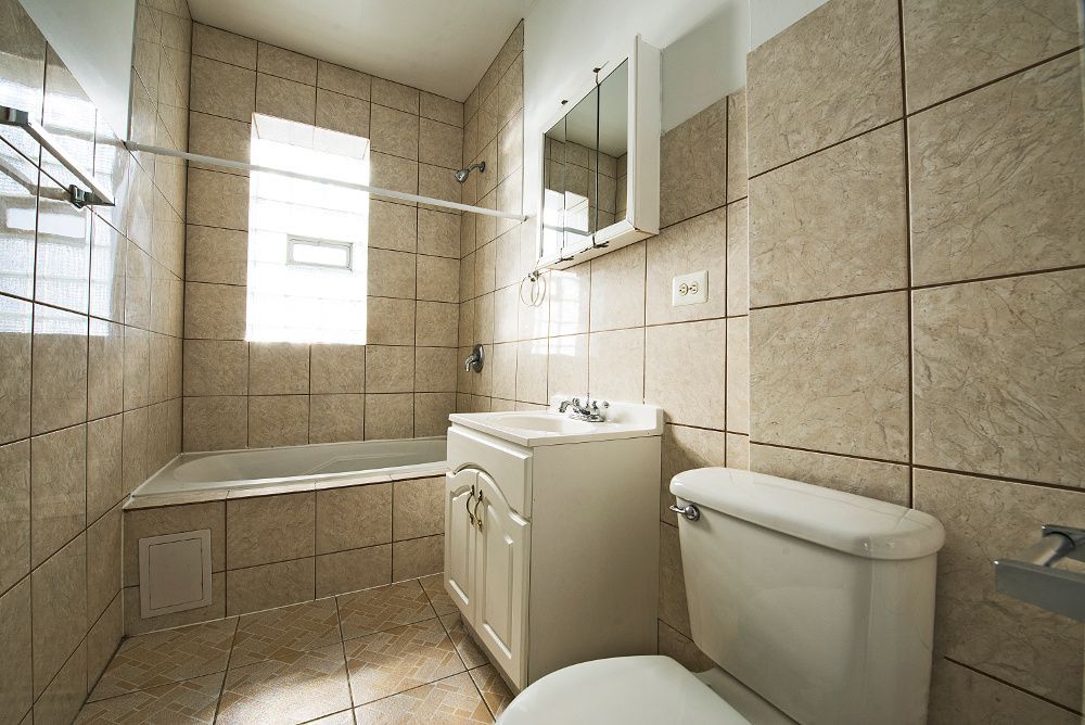 Bathroom with beige tiled walls and floor, white fixtures: sink, toilet, tub, and window.