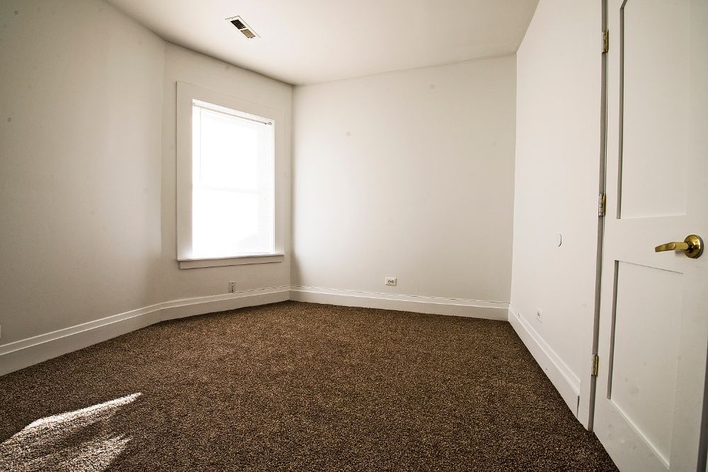 Empty room with brown carpet, white walls, a window, and a closed white door.