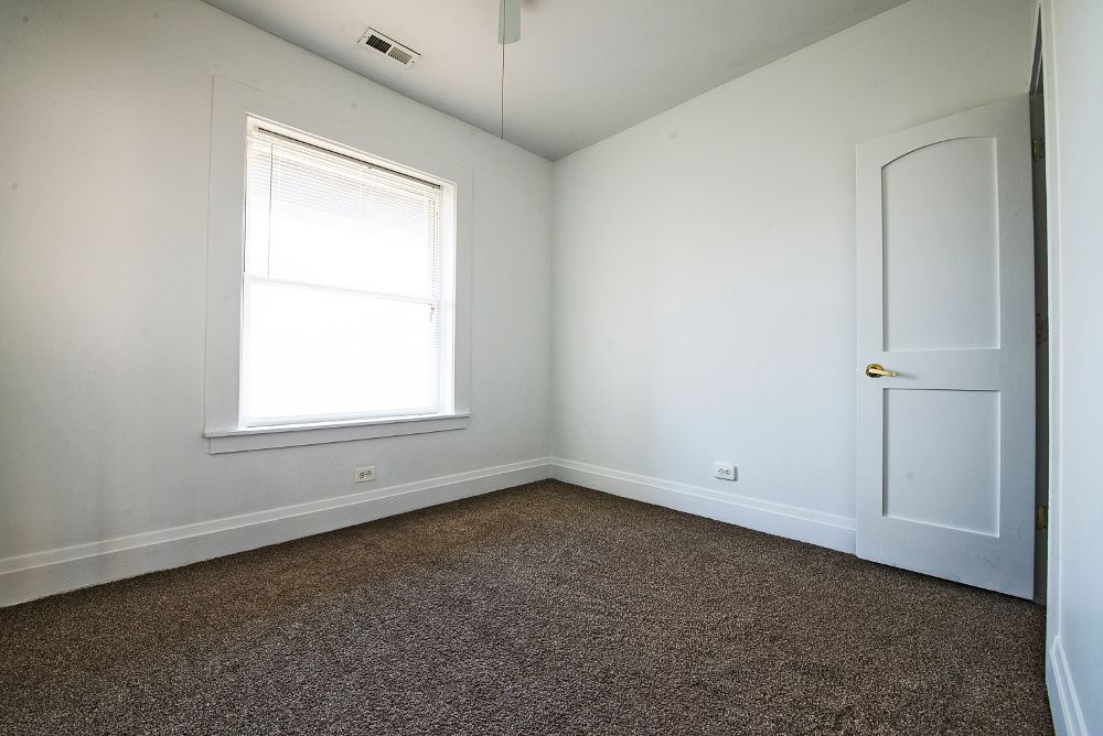 Empty, light-filled room with brown carpet, white walls, window, and door.