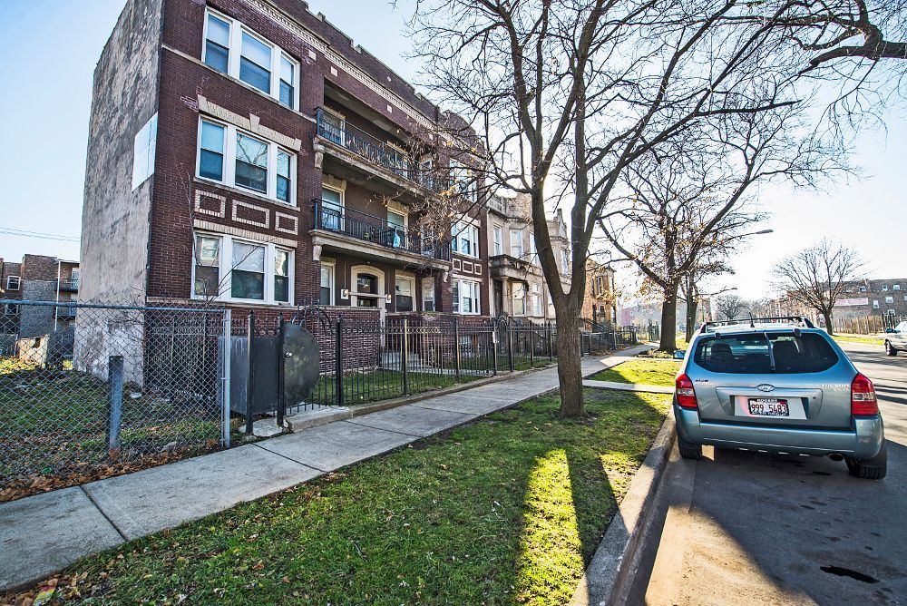Brick apartment building with a sidewalk, grassy area, fence, and parked silver car.