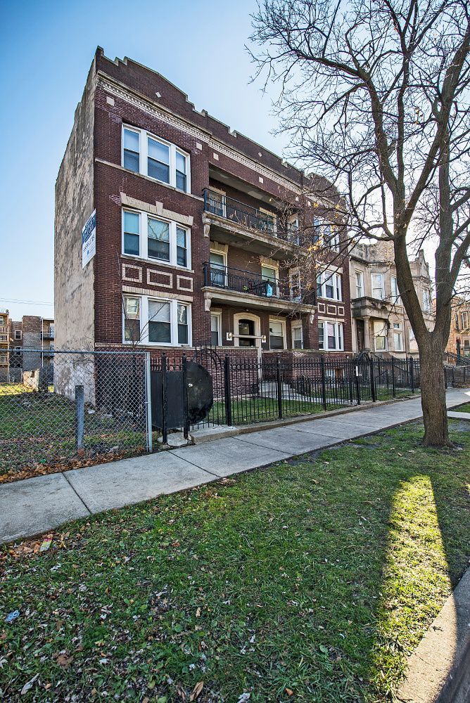 Brick apartment building with balconies, sidewalk, grass, and a chain-link fence.