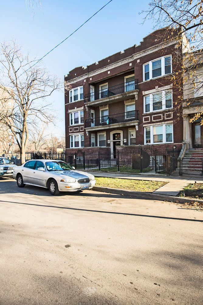 A light-colored sedan driving on a city street in front of a brick apartment building.