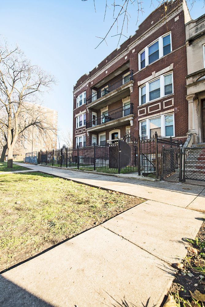 Three-story brick building with balconies behind a wrought-iron fence on a sunny day.