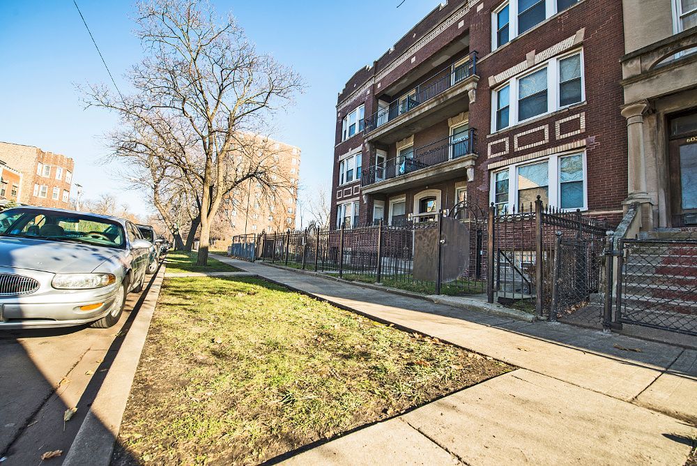 Apartment building on a sunny street with a car parked nearby and a small yard with a fence.