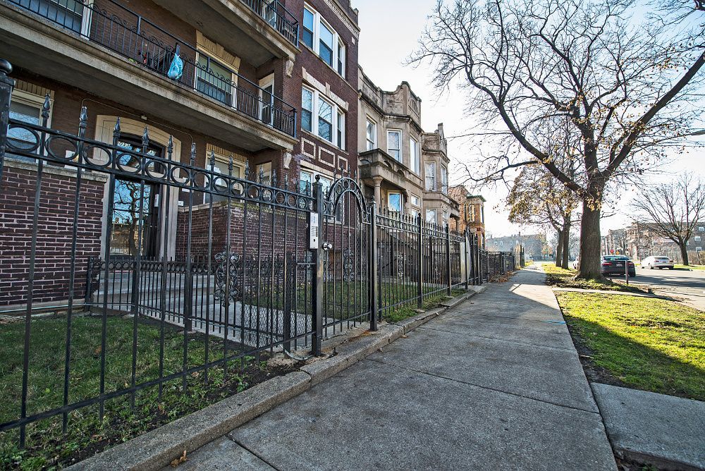 Apartment buildings behind a black fence, on a sunny day. Sidewalk and bare tree on the right.