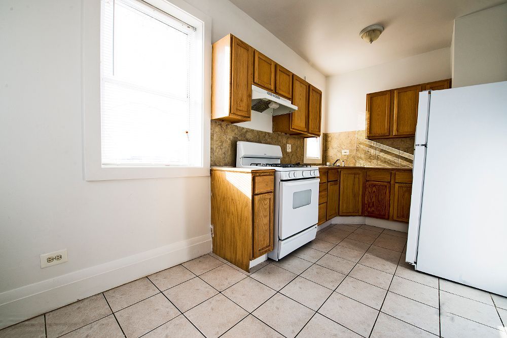 Kitchen with wooden cabinets, white appliances, and tiled floor. Light streams through the window.