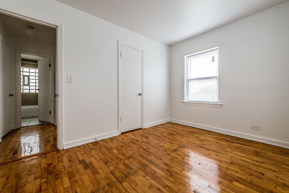 Empty room with hardwood floors, white walls, closed door, and window. Another room visible through doorway.