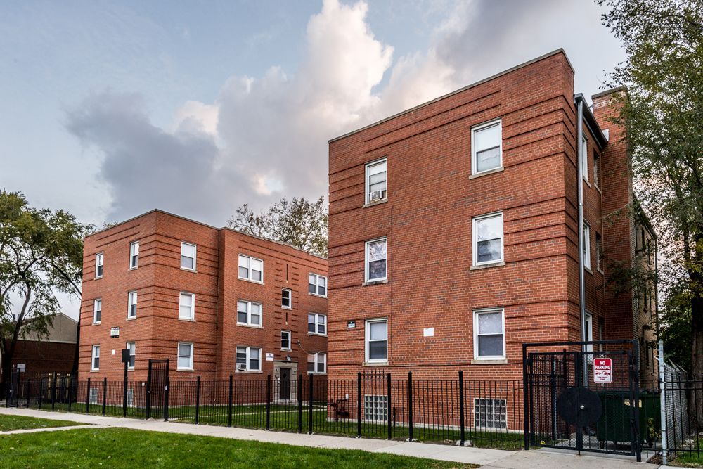 Two red brick apartment buildings behind a black metal fence, under a cloudy sky.