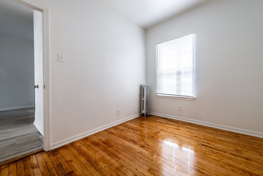 Empty room with hardwood floors, white walls, small window with blinds, and a radiator.