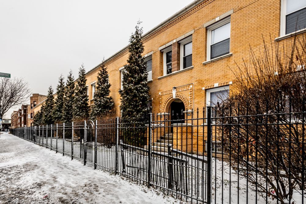 Row of tan brick apartment buildings behind a black metal fence, snowy sidewalk.