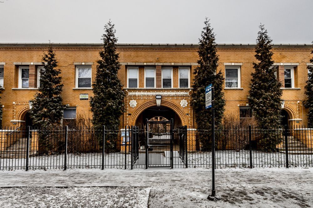 Yellow brick building with archway entrance, black iron fence, snow-covered ground, evergreen trees.