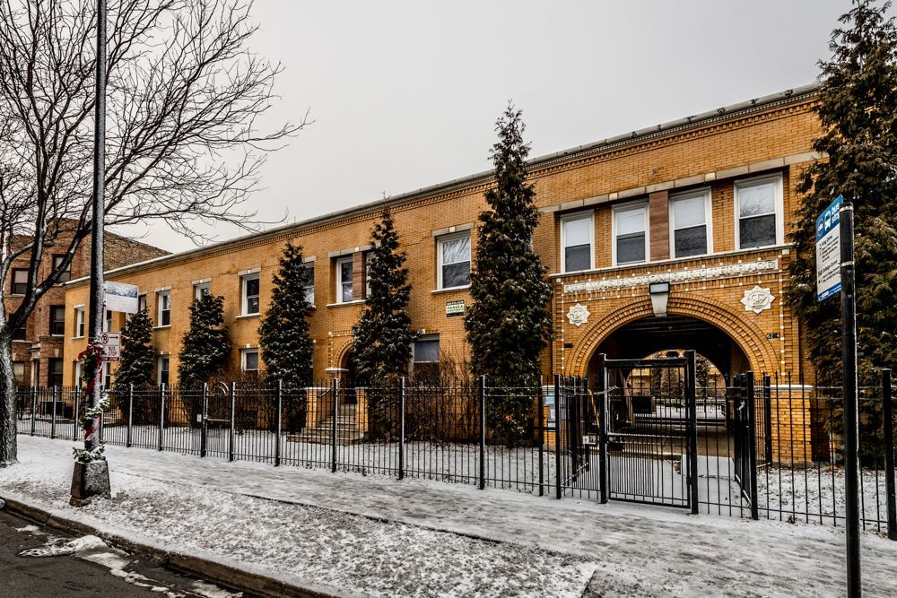 Brick building with arched entrance and snow-covered ground; trees and wrought-iron fence.