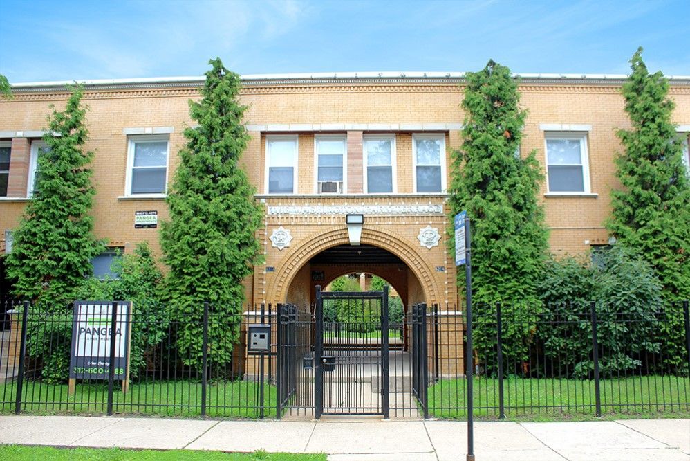 Two-story brick building with arched entrance and black wrought-iron gate, flanked by tall green trees.
