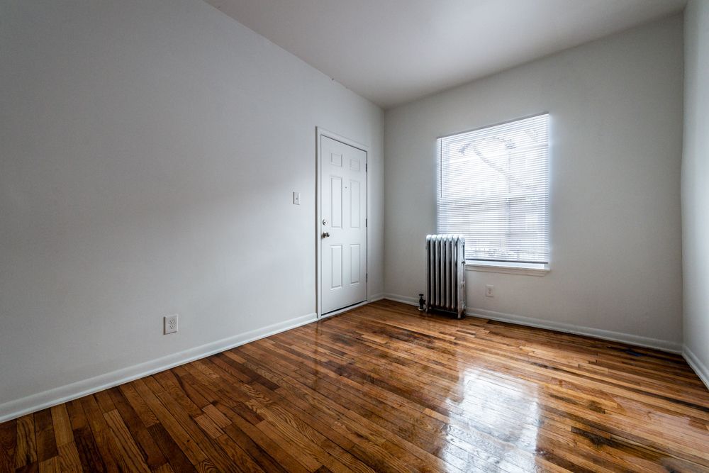 Empty room with hardwood floors, white walls, door, window with blinds, and radiator.