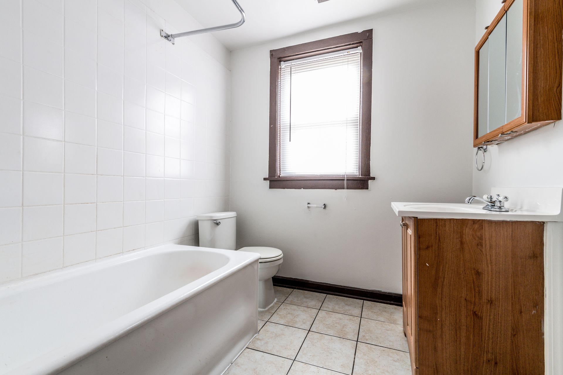 Bathroom with white tiled walls, a bathtub, toilet, window, and wood vanity.