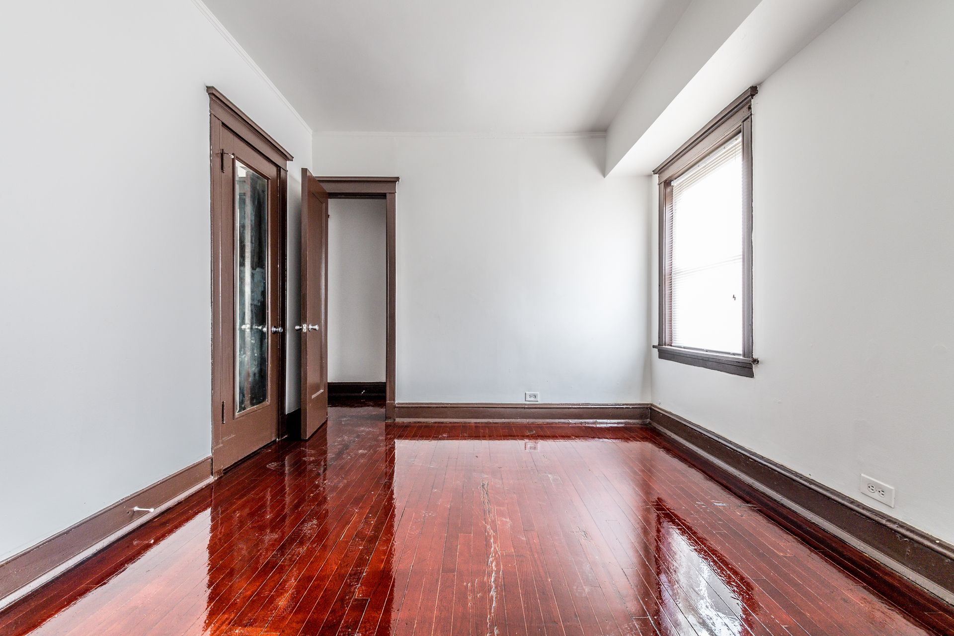 Empty room with glossy hardwood floors, white walls, dark wooden trim, and window.