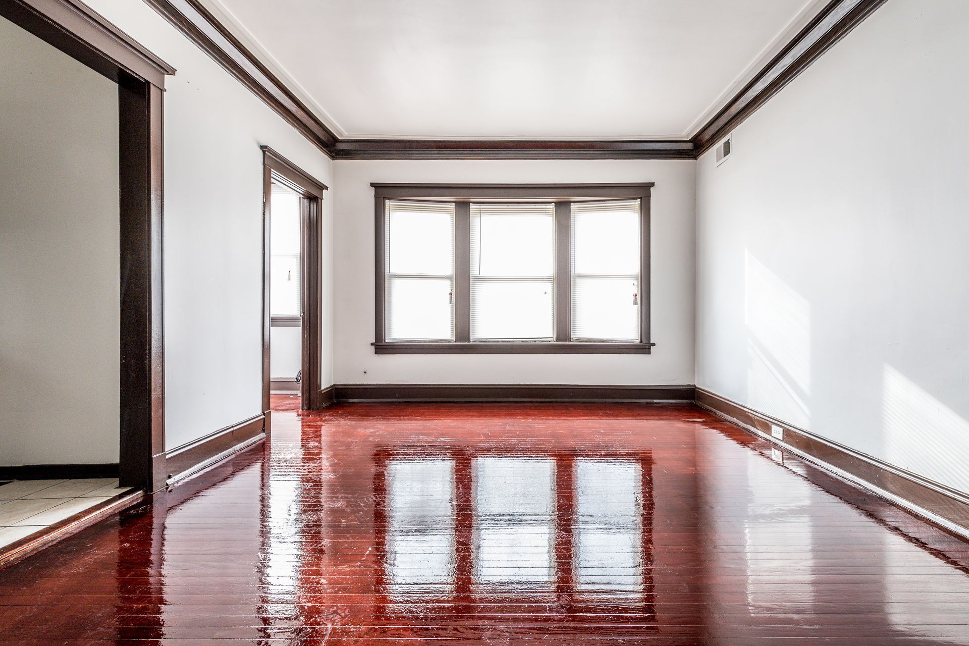 Empty room with shiny, red-brown floors, white walls, and dark wood trim around a window and doorway.