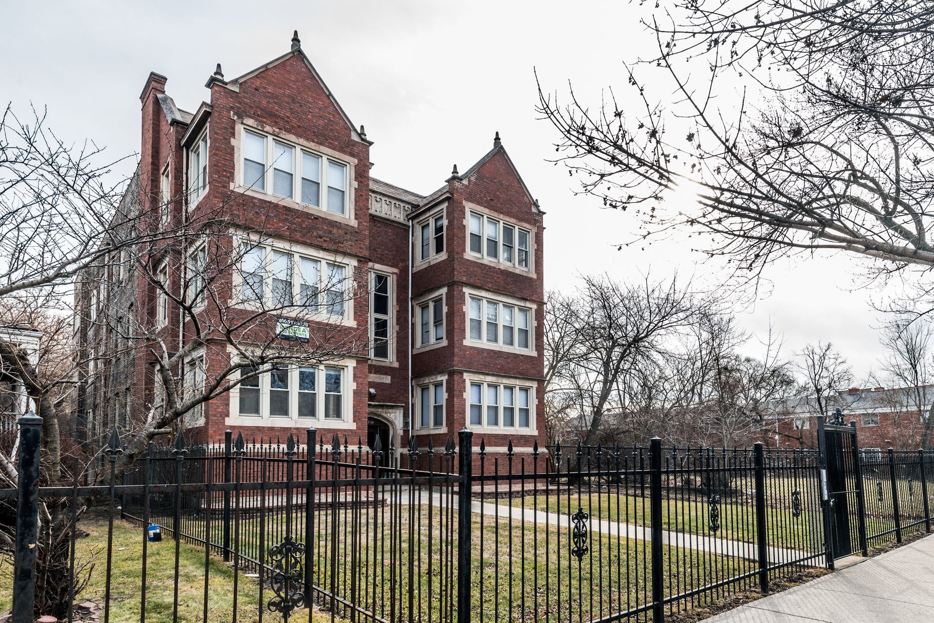 Brick apartment building with black fence in front. Bare trees and cloudy sky.