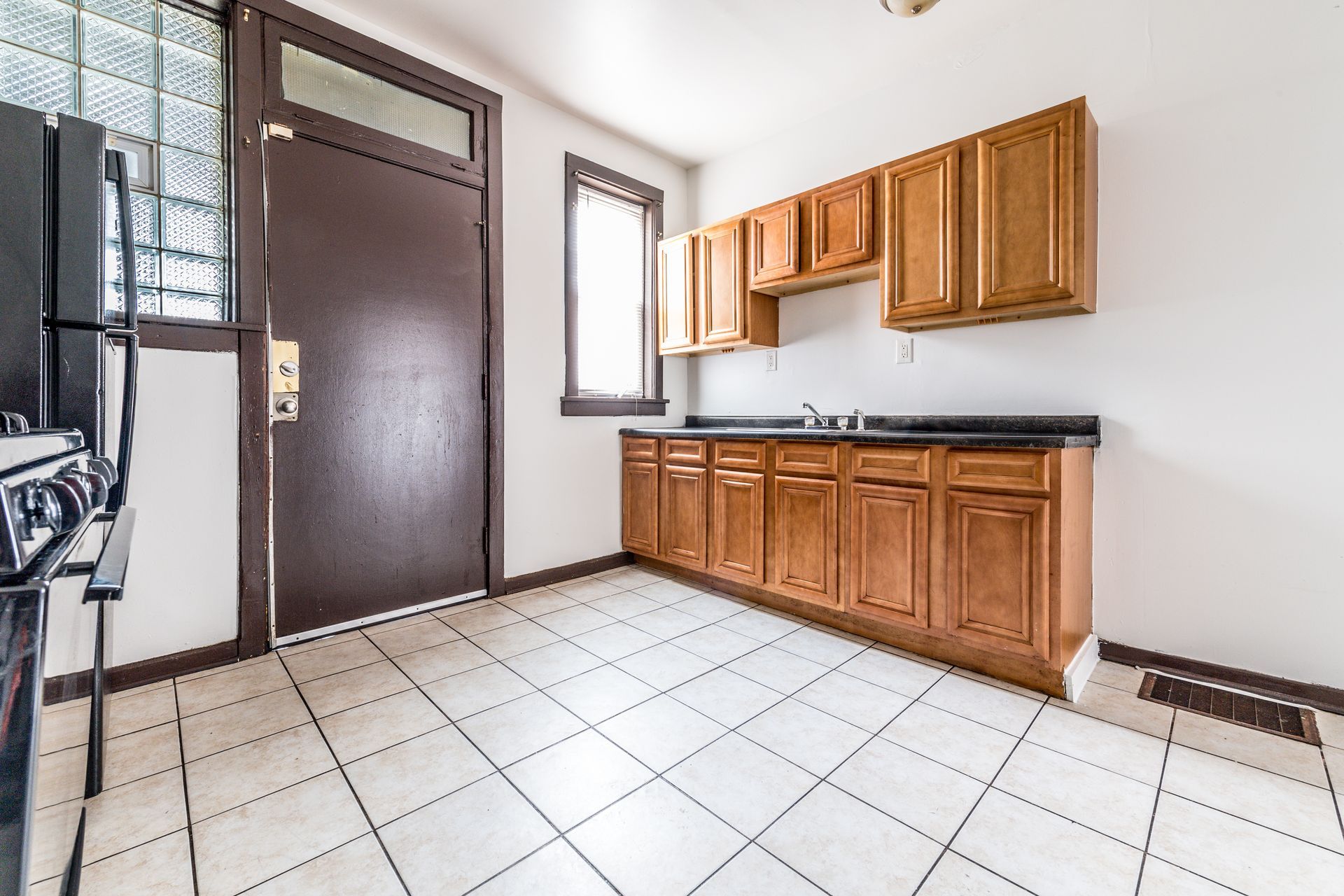 Kitchen with wooden cabinets, black countertop, white tile floor, brown door, and small window.