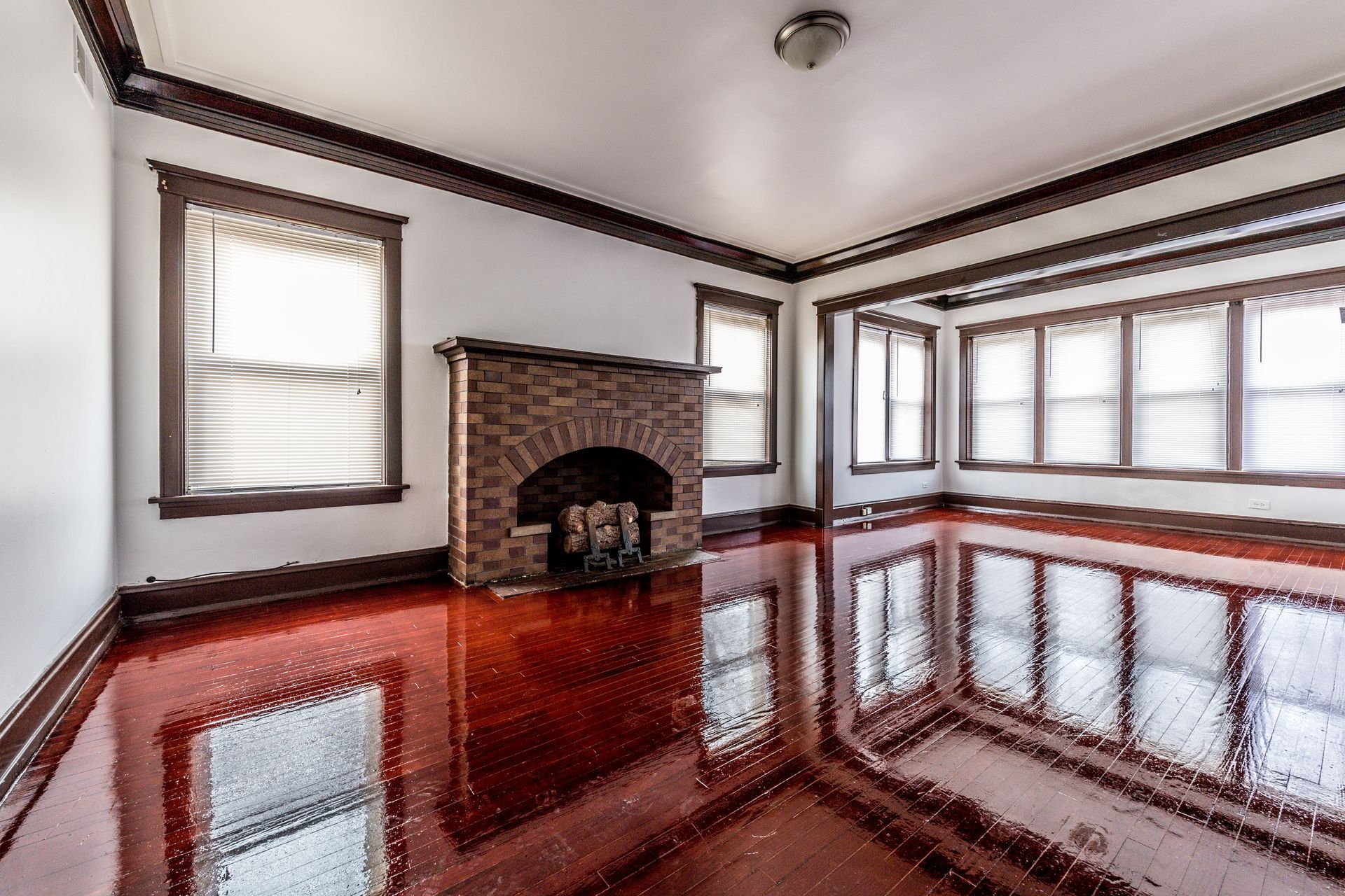 Empty room with shiny, red floor, brick fireplace, and dark wood trim. Windows provide natural light.