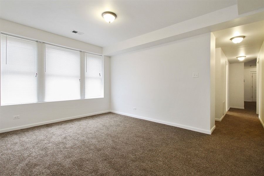 Empty room with brown carpet, white walls, and a hallway. Windows with blinds.
