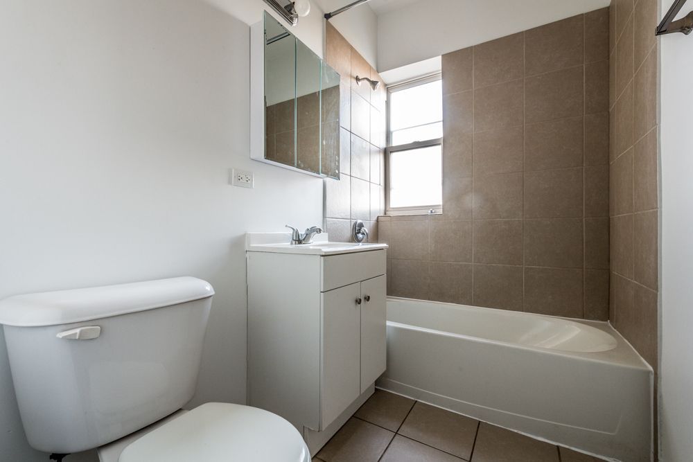 Bathroom with white toilet, vanity, and bathtub. Beige tiled wall and window.