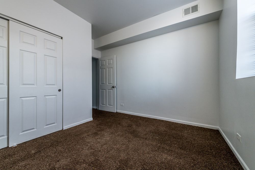 Empty bedroom with brown carpet, white walls, and closet. A door leads to another room.