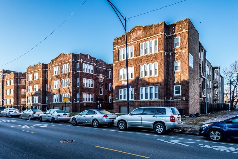 Brick apartment buildings along a street with parked cars on a sunny day.