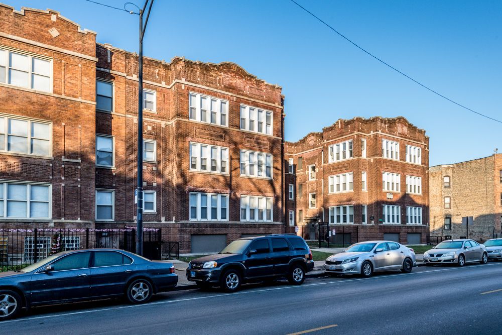 Brick apartment buildings line a street with parked cars on a sunny day.