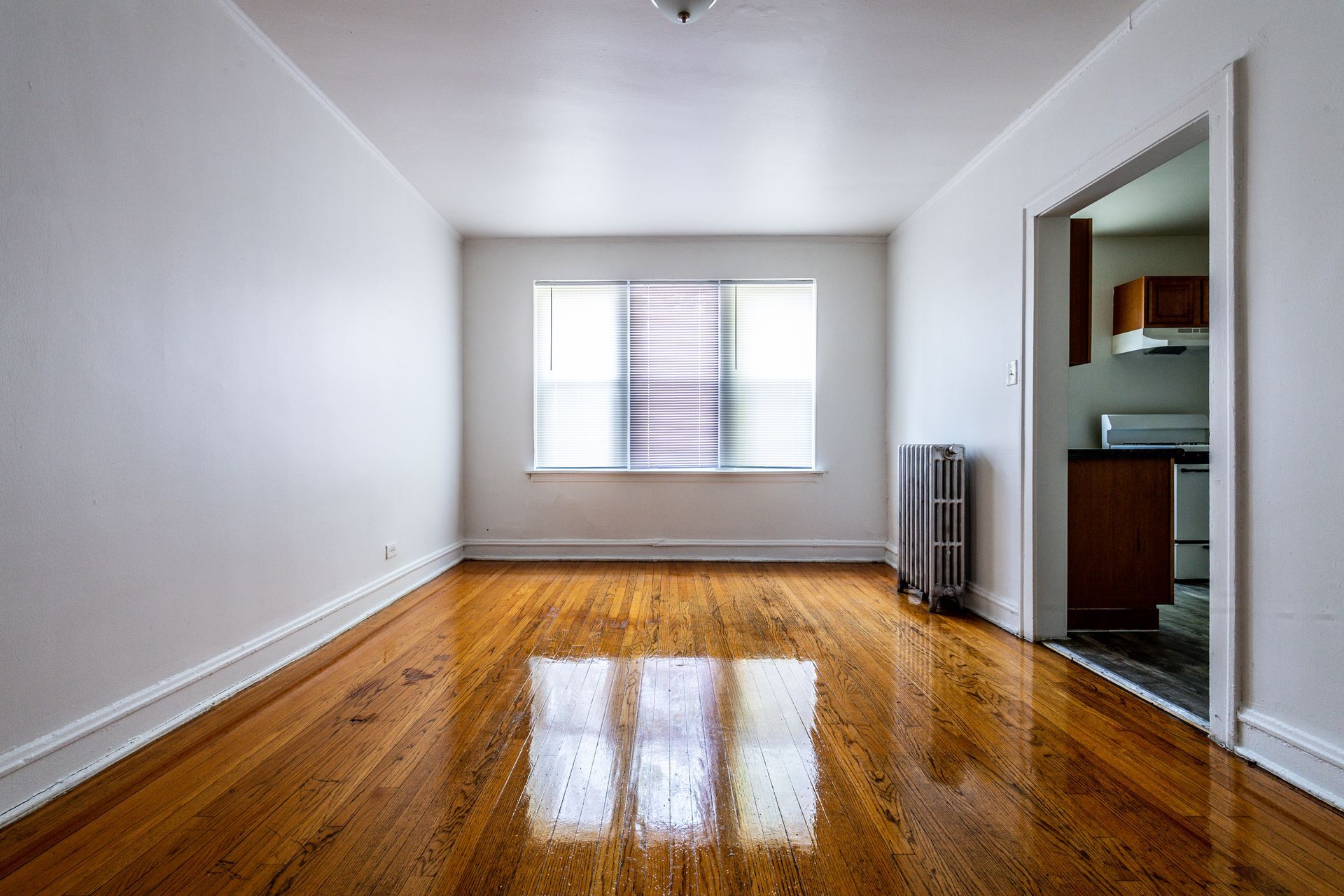 Empty room with hardwood floors, a window, and an open doorway to a kitchen.