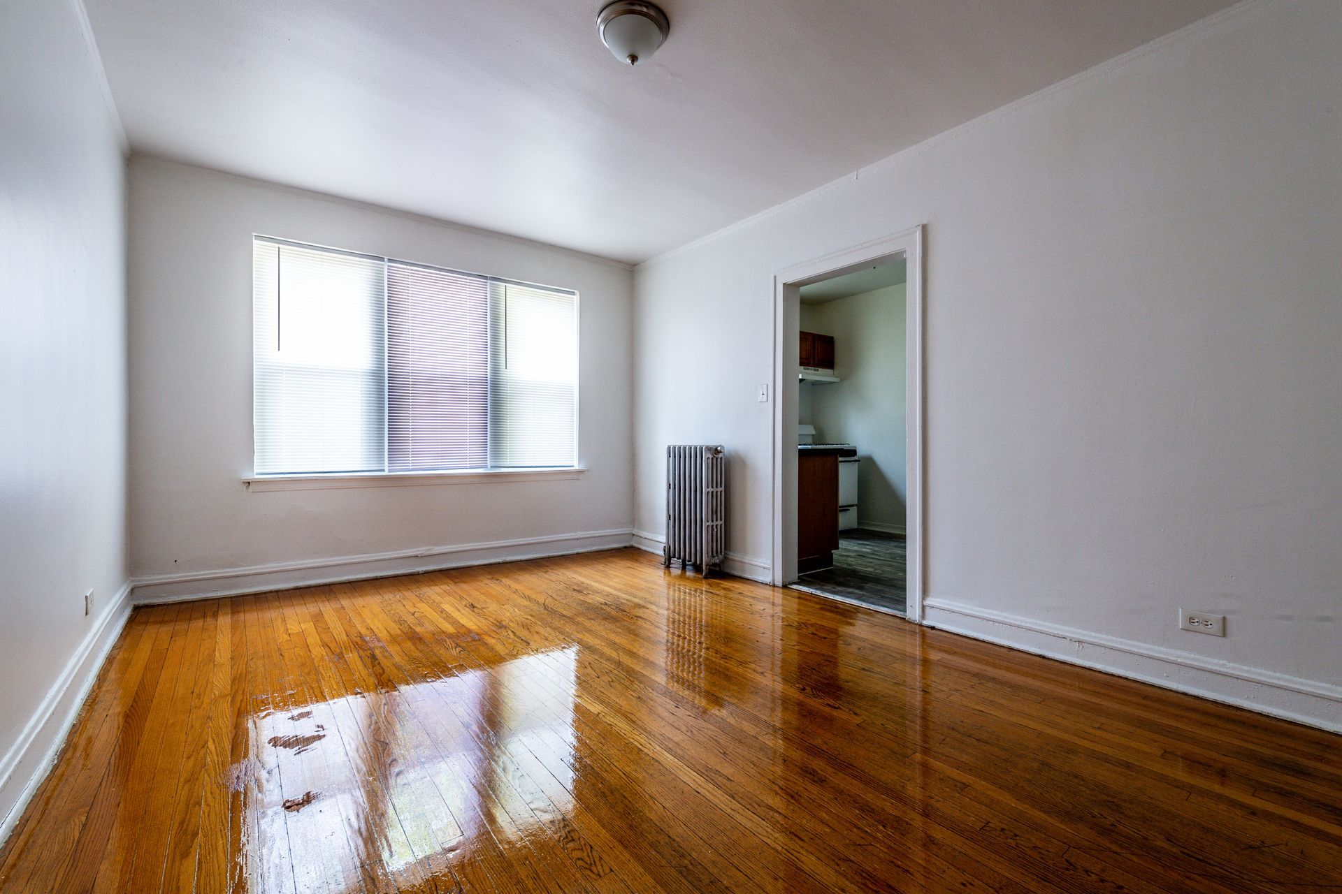 Empty room with hardwood floors, window with blinds, and doorway to a kitchen.