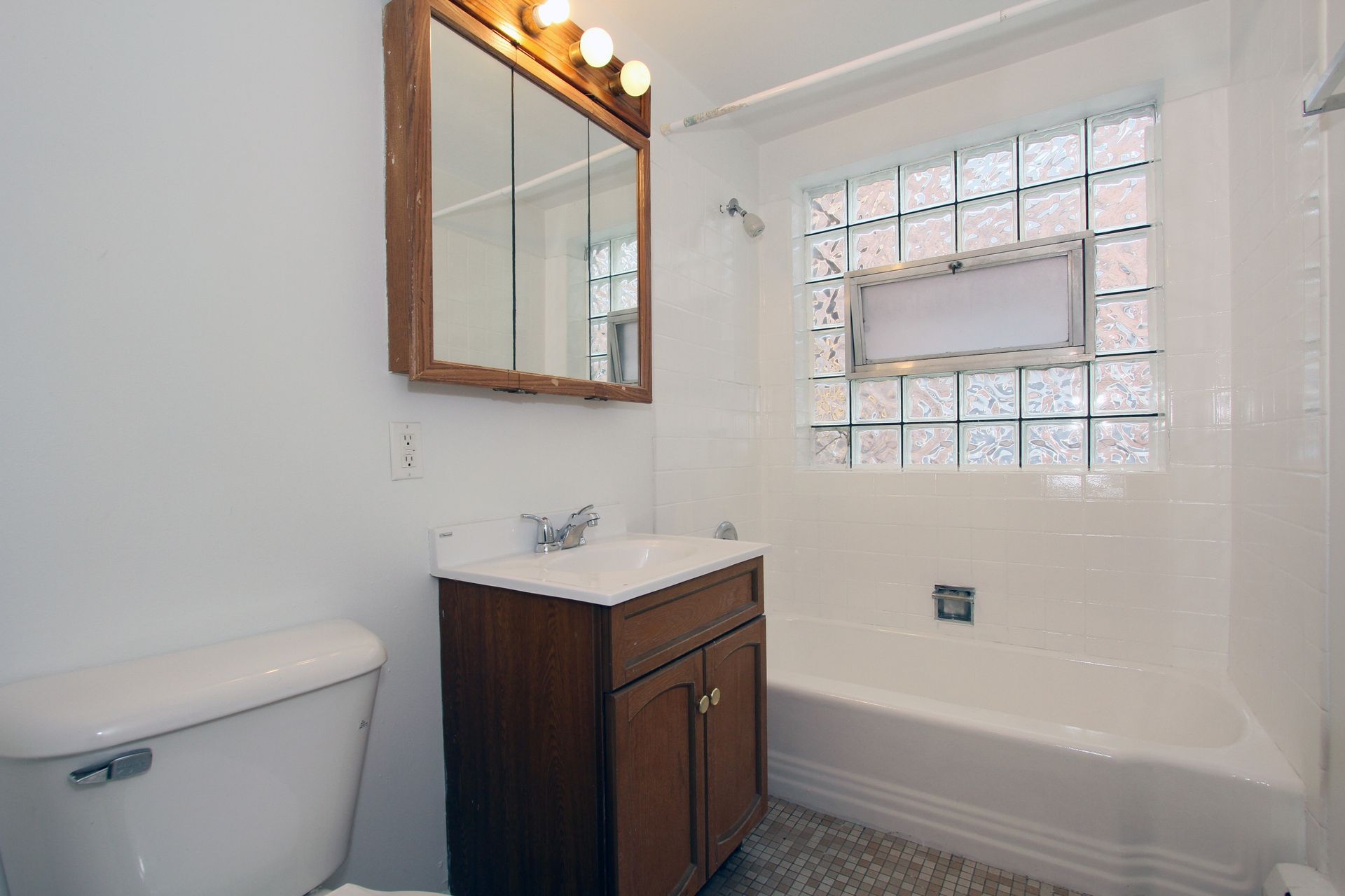 Bathroom with white walls, a brown vanity, and a glass block window above a bathtub.