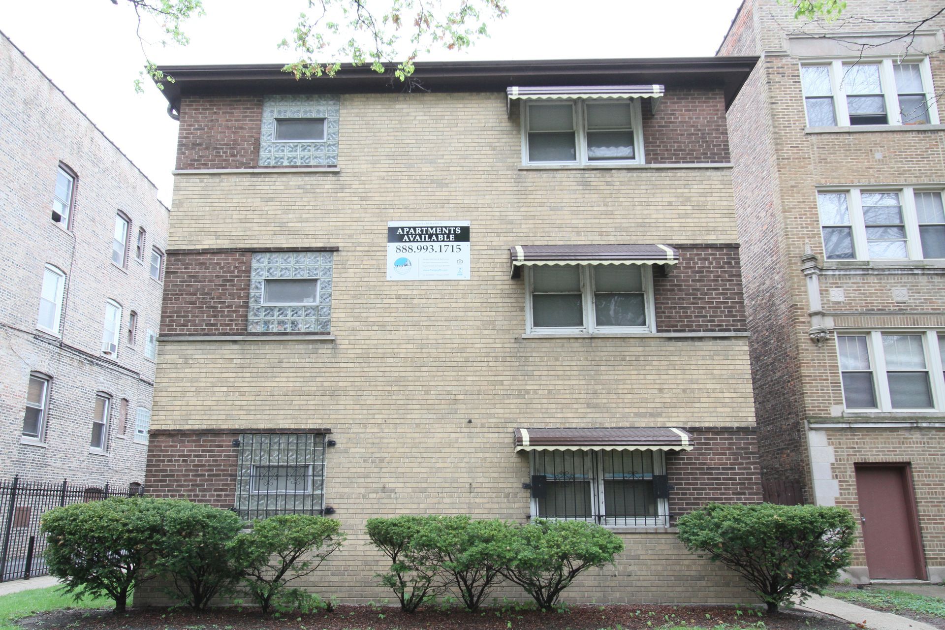 Three-story brick apartment building with awnings over windows. Green bushes in front.
