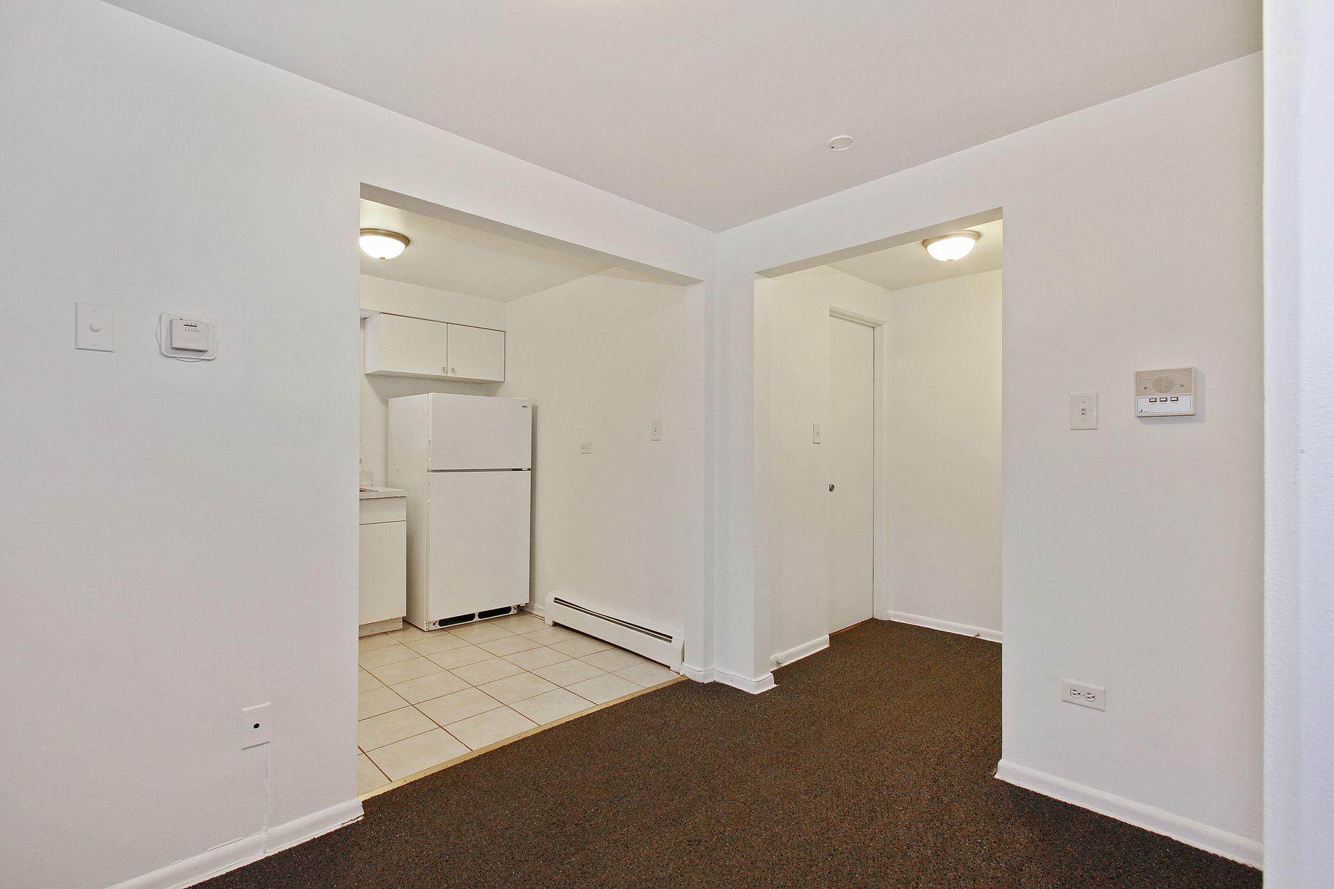 Empty apartment interior with a glimpse of the kitchen and a doorway. Dark carpet, white walls, and a tiled floor.