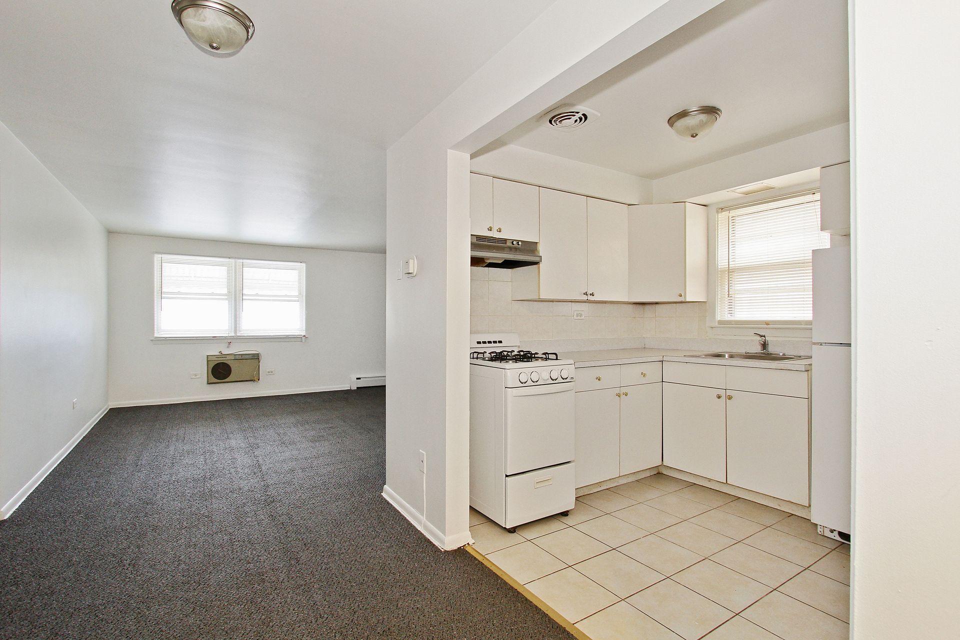 Empty apartment interior with kitchen: white cabinets, stove, and a window; carpeted living area.