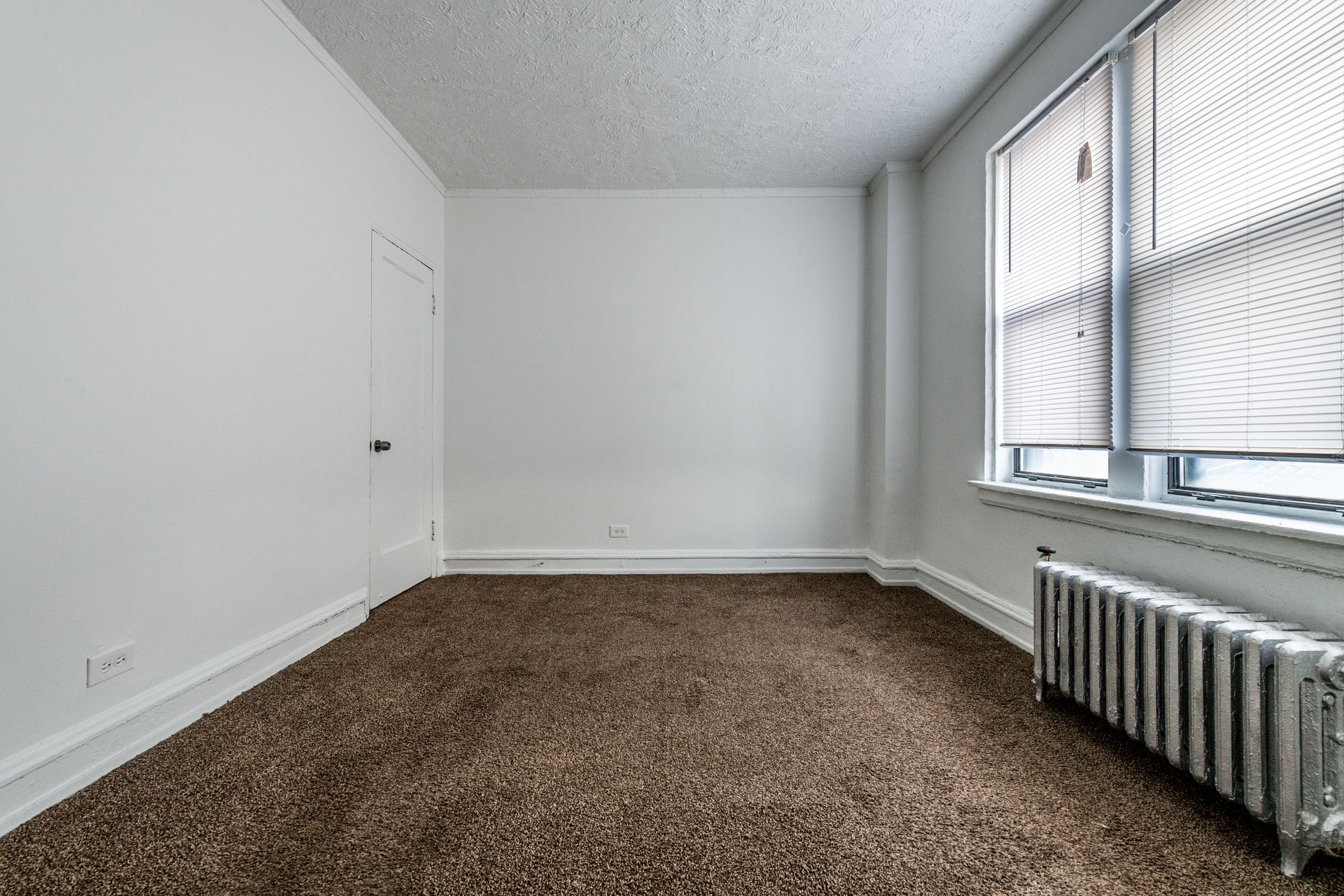 Empty room with brown carpet, white walls, small door, window with blinds, and radiator.