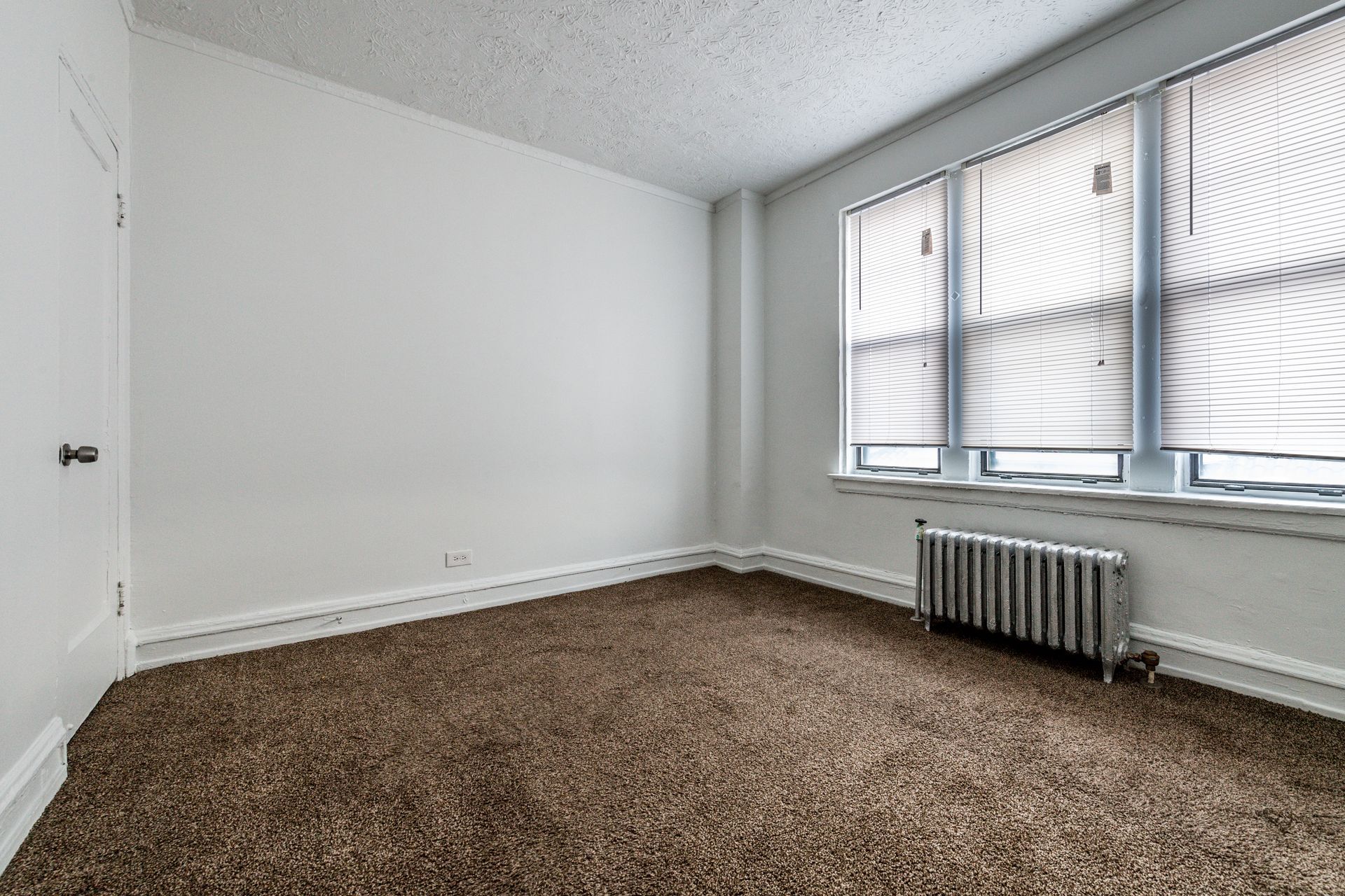 Empty room with brown carpet, white walls, and a radiator under a window with blinds.