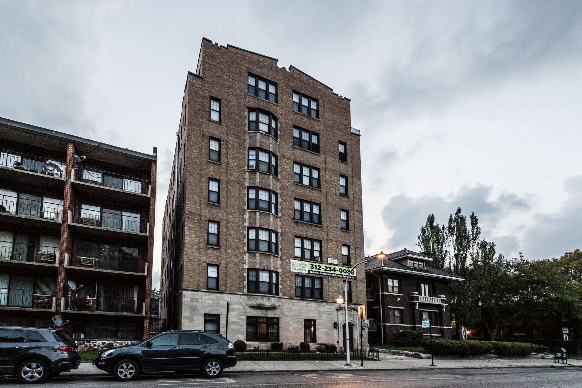 Tall brick apartment building on a city street, overcast sky. Black cars parked in front, trees on right.