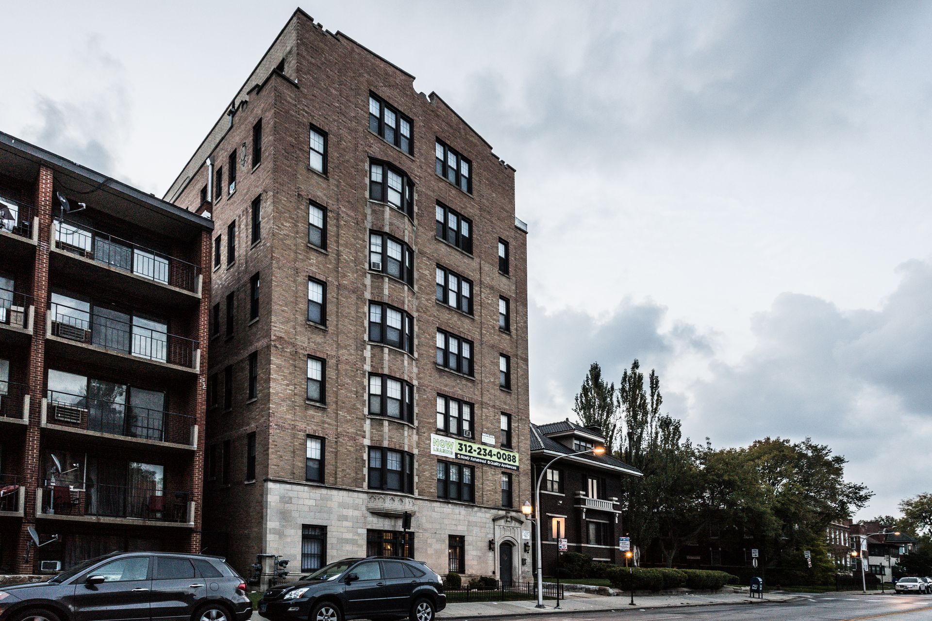 Brick apartment building on a city street under cloudy skies, cars parked in front.
