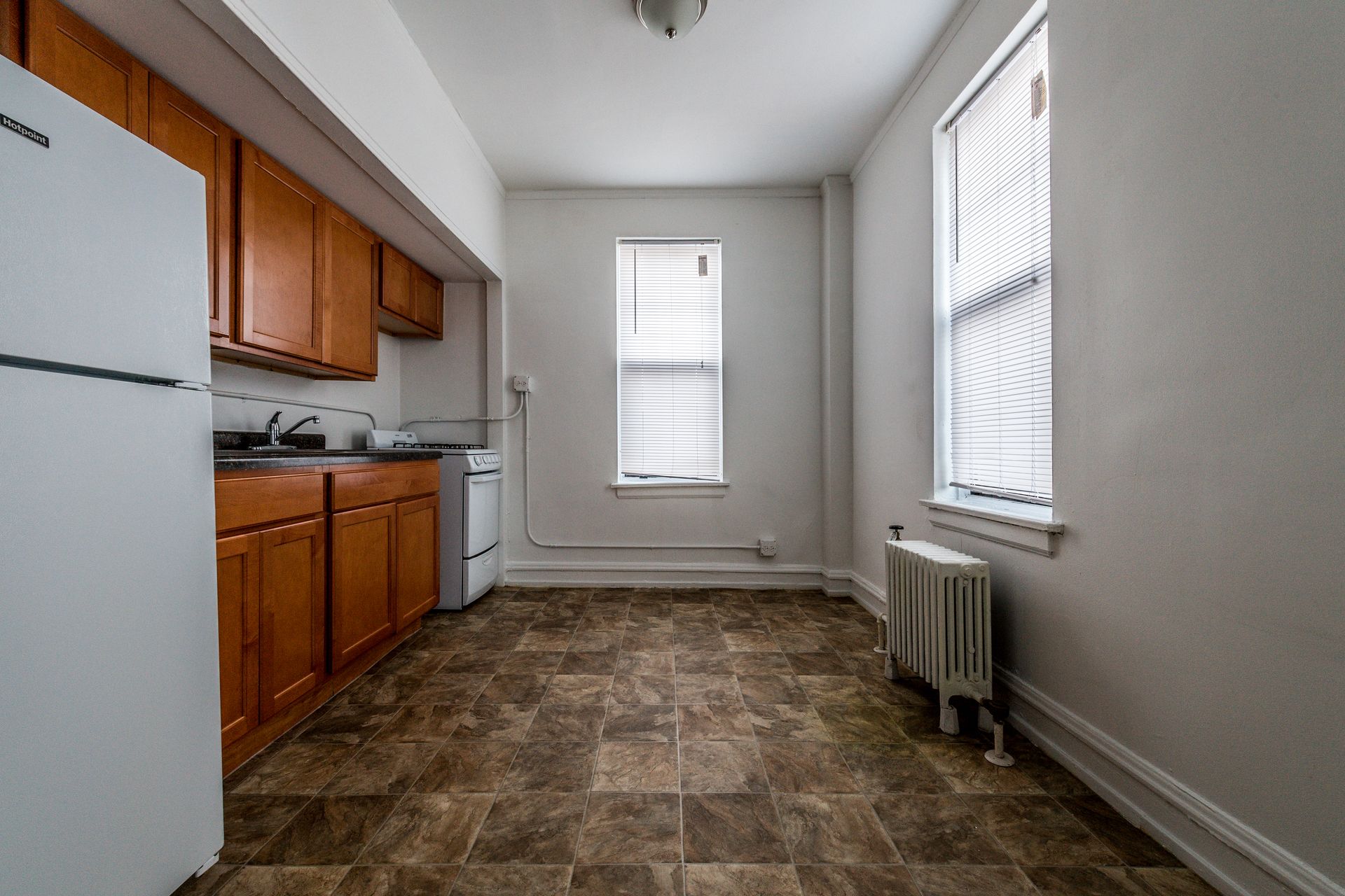 Kitchen with brown cabinets, white appliances, and tiled floor; two windows with blinds.