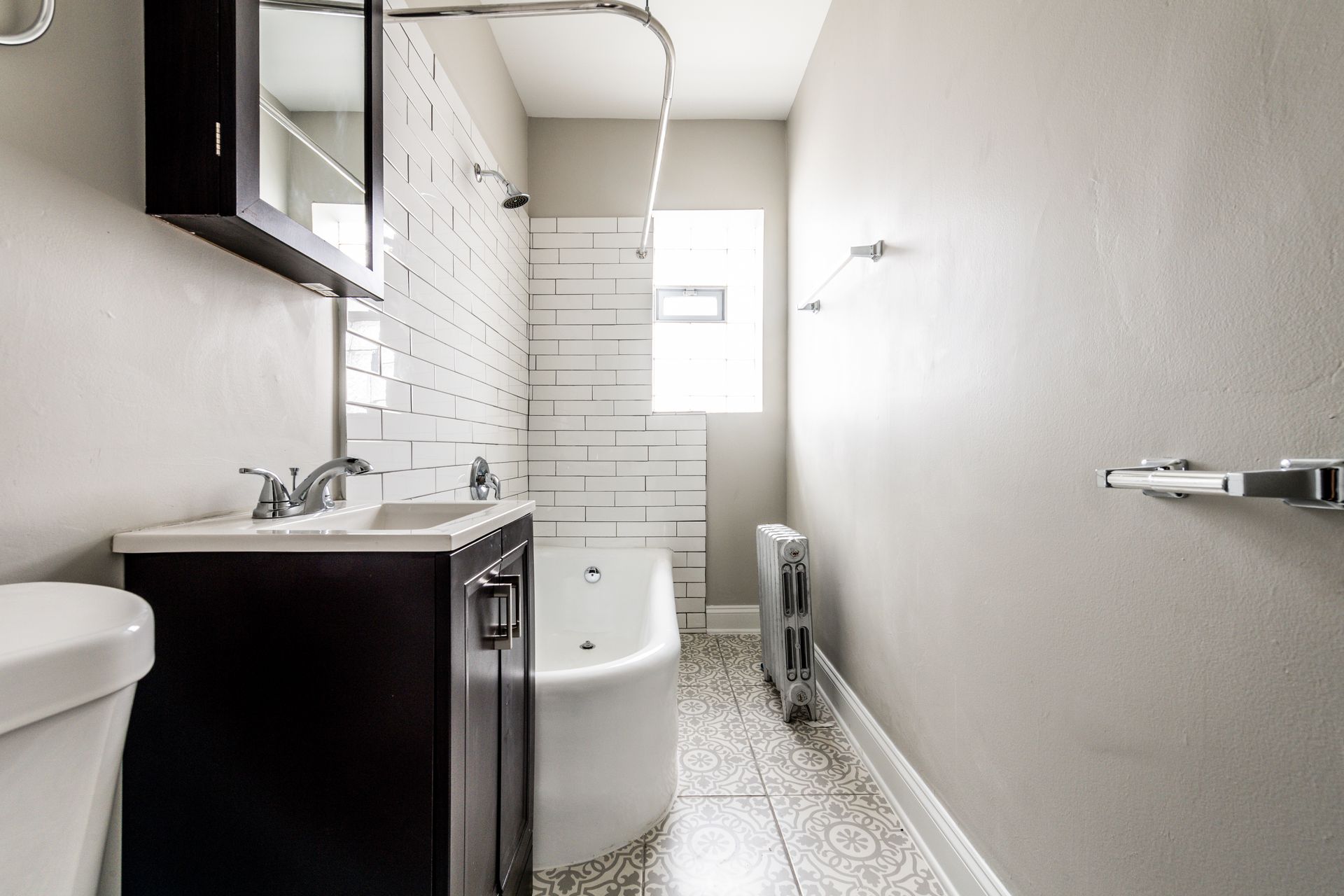 Bathroom with white tile, dark vanity, and gray walls. Includes a tub, toilet, and radiator.