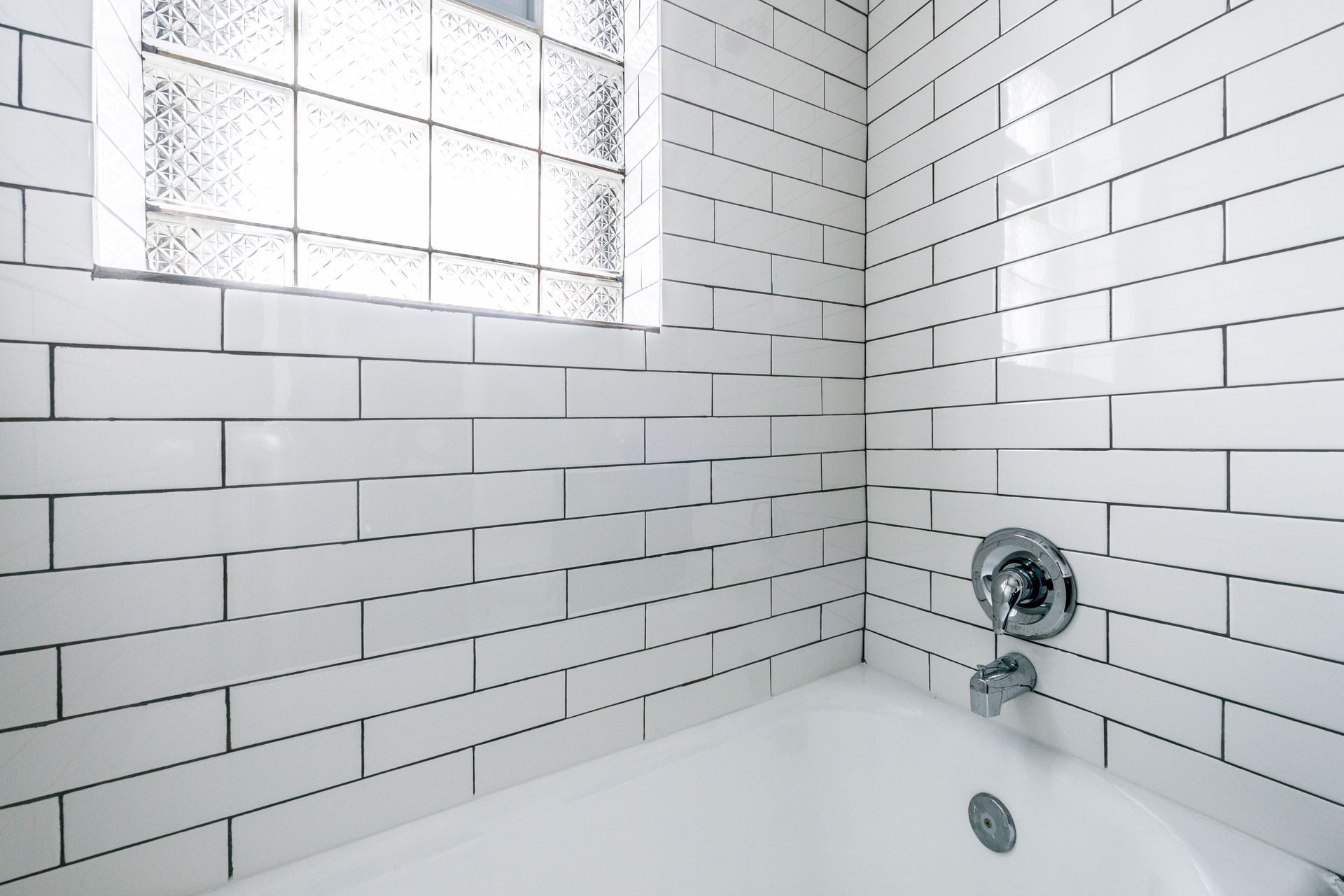 White tiled bathroom with tub, shower fixture, and frosted glass window.