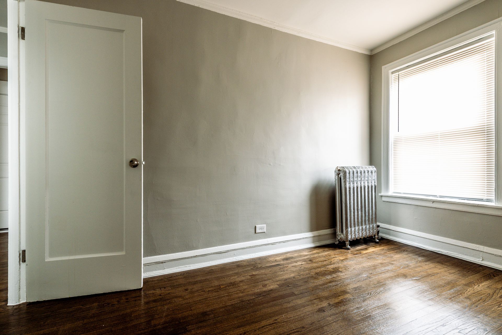 Empty room with gray walls, wood floor, white door and trim, window with blinds.
