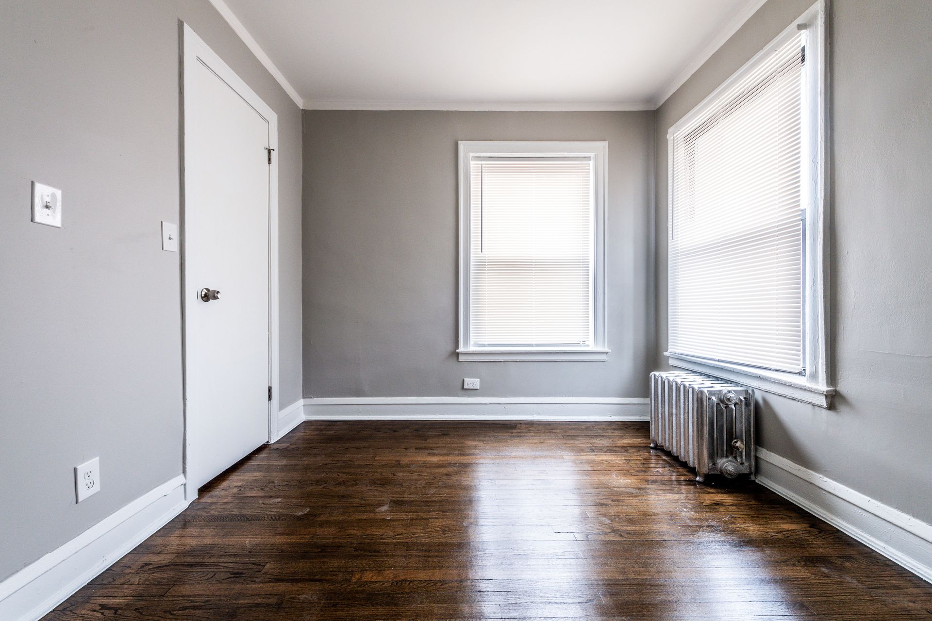 Empty room with gray walls, wood floor, white door, and window with blinds.