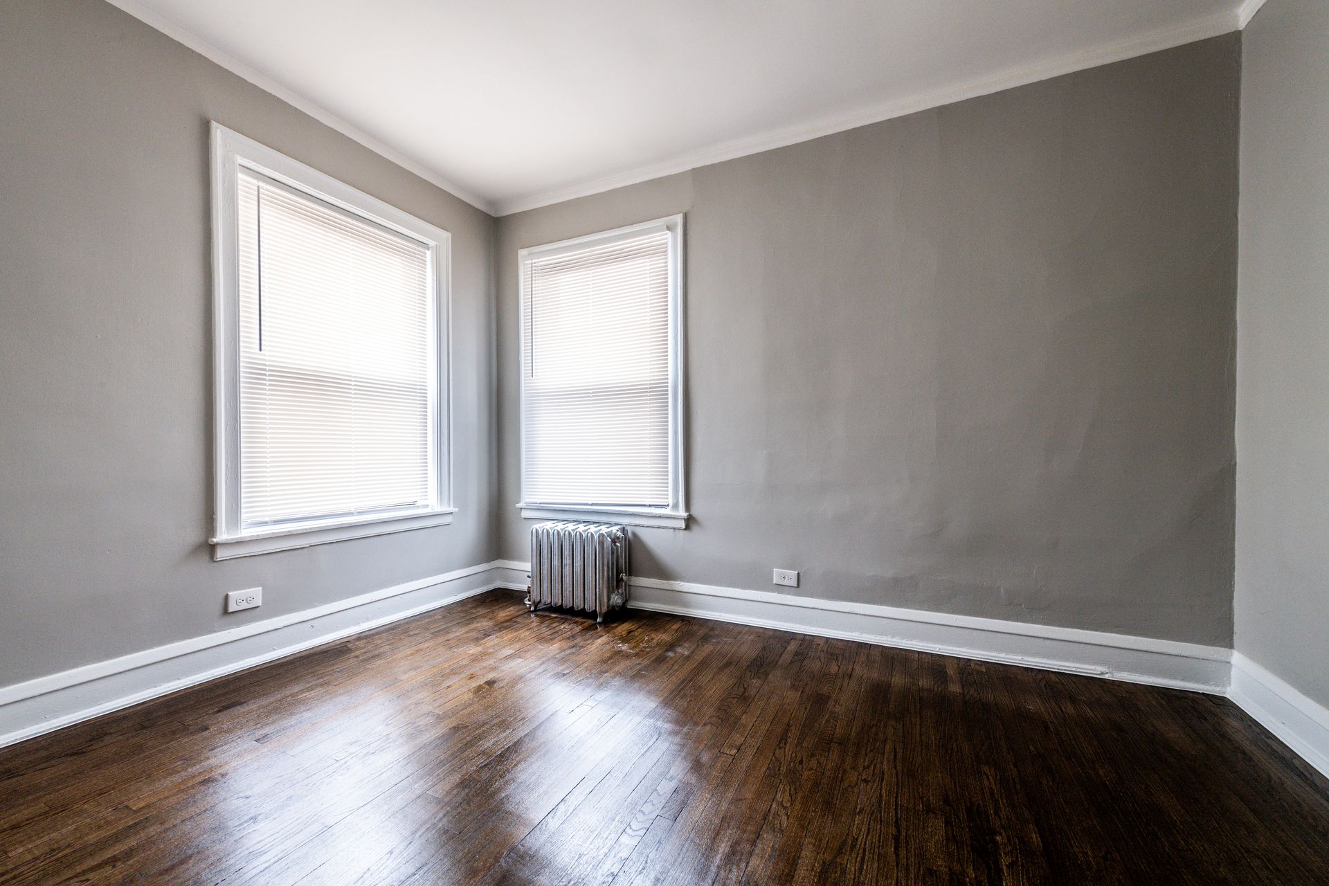 Empty room with gray walls, two windows with blinds, dark wood floor, and radiator.