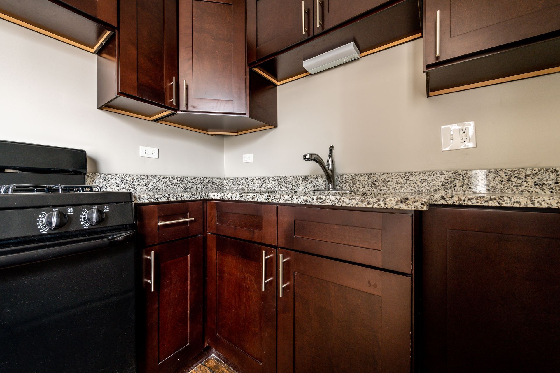 Dark wood kitchen cabinets, black stove, granite countertop, stainless faucet, and a light-colored wall.