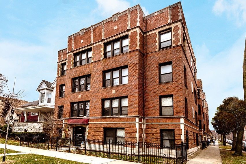 Four-story brick apartment building with dark trim, sidewalk, and a small fence.
